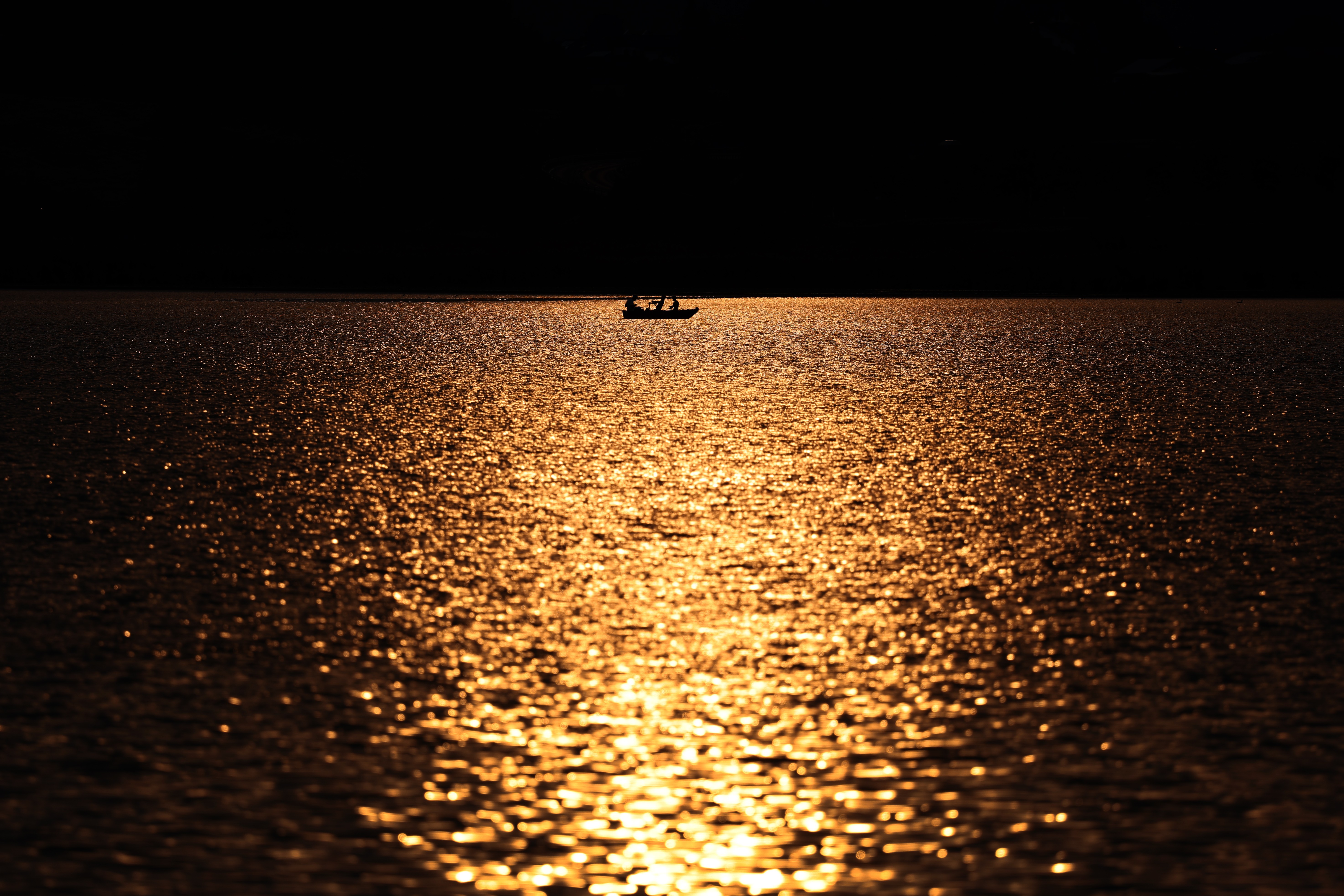 Anglers row a boat across a lake that glistens in the sunset.