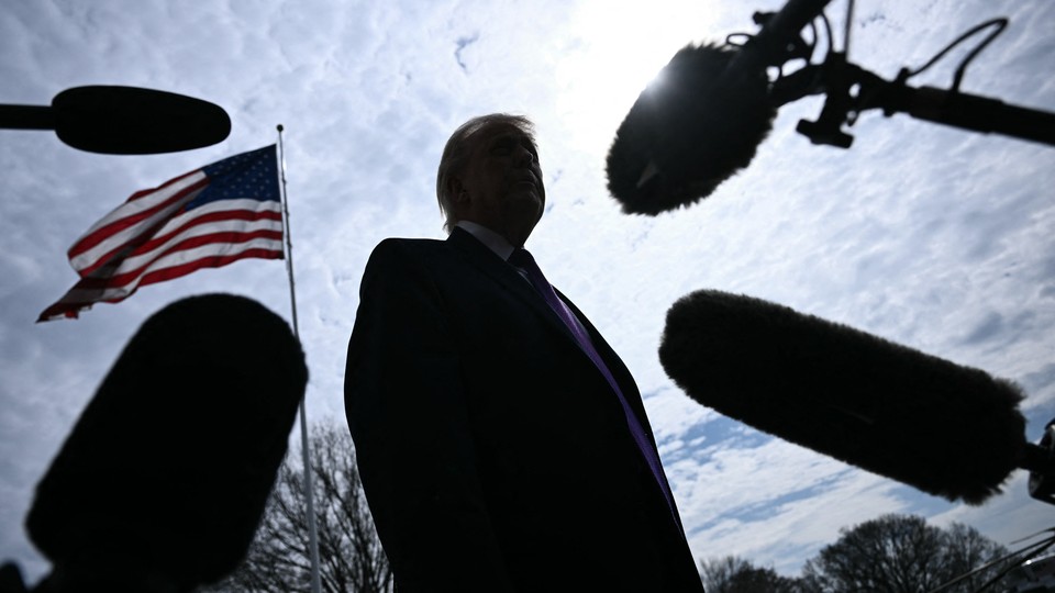An image of Trump's silhouette from below surrounded by microphones, with an American flag flying in the background.
