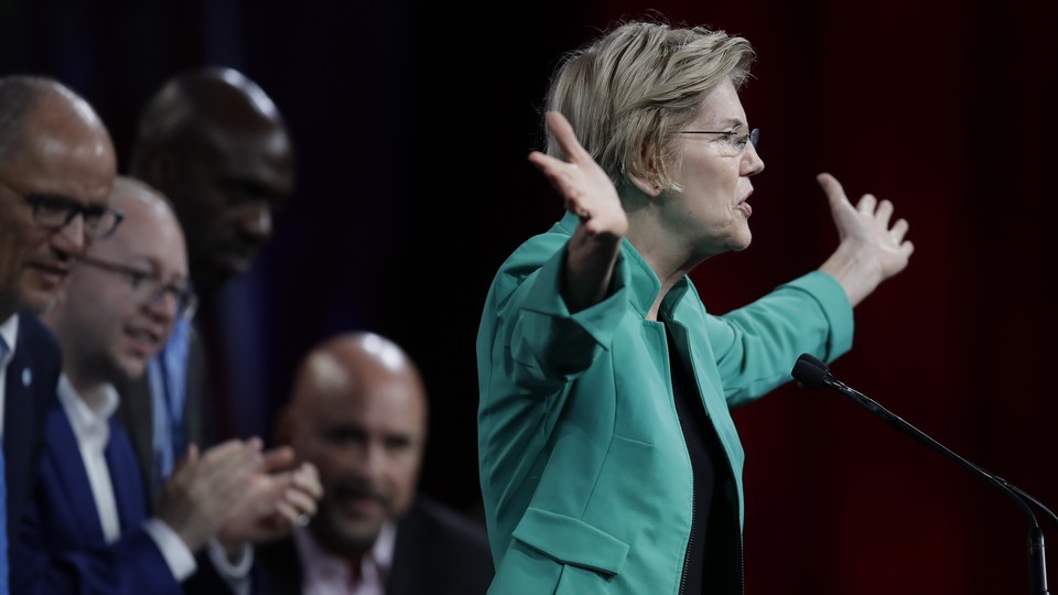 Elizabeth Warren, dressed in an open green jacket, stretches her arms out to her sides as she speaks to an audience. Three men are visible onstage behind her.