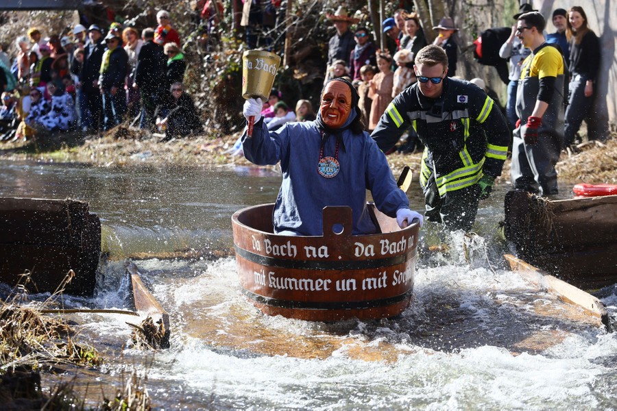 A person wearing a mask, sitting in a tub, floats down a stream as a crowd looks on.