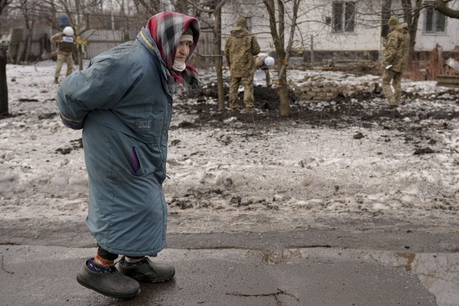 A woman wearing along winter coat walks past several people wearing camouflage gear who are inspecting an artillery crater.