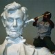 A person uses a cloth-wrapped pole to clean the ear of a large statue of Abraham Lincoln.