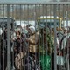 A line of people bundled up in parkas and scarves wait in a crowded line behind a tall barred metal fence with buses and snowy city in background