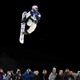 A crowd looks up as a snowboarder makes a high jump in the dark sky above them.