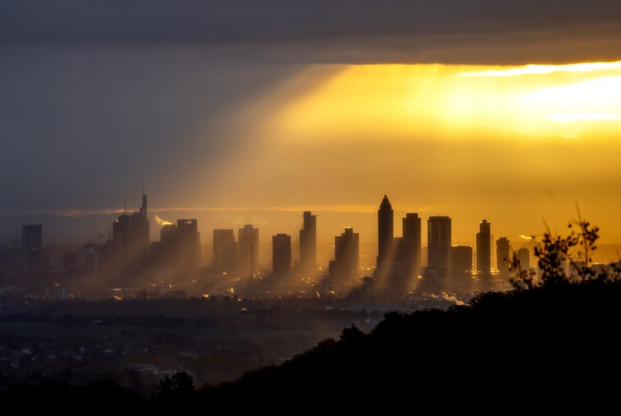 Sunshine pours from a hole in a cloudy sky, streaming between distant tall buildings.