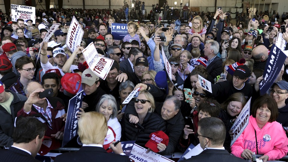 A crowd of people, many holding Trump signs, clamor to get Trump's attention, who is standing at the front of the crowd signing a hat.