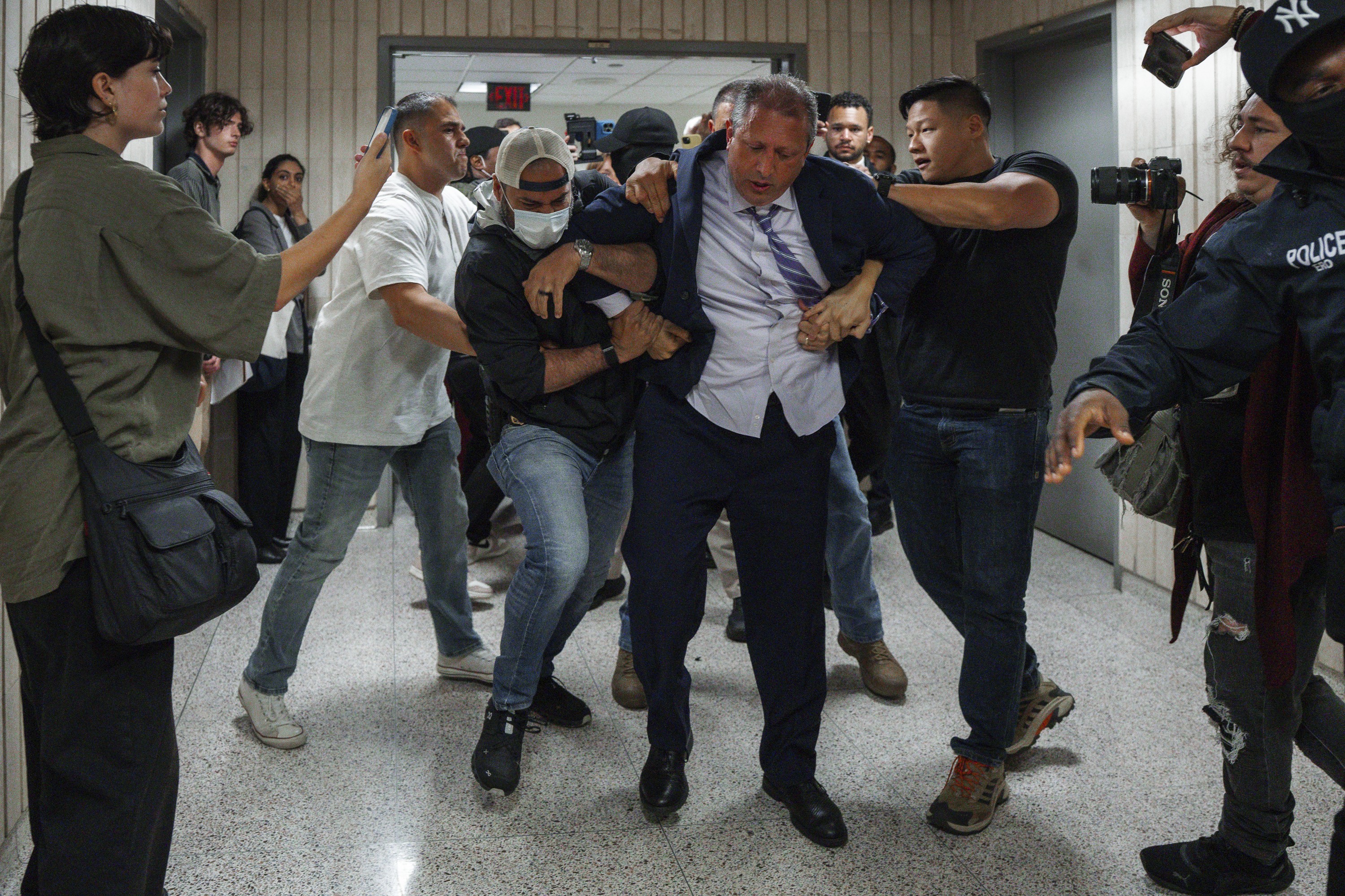Several men in civilian clothing struggle with a man wearing a suit, aggressively escorting him down a hallway past people taking photos and videos.