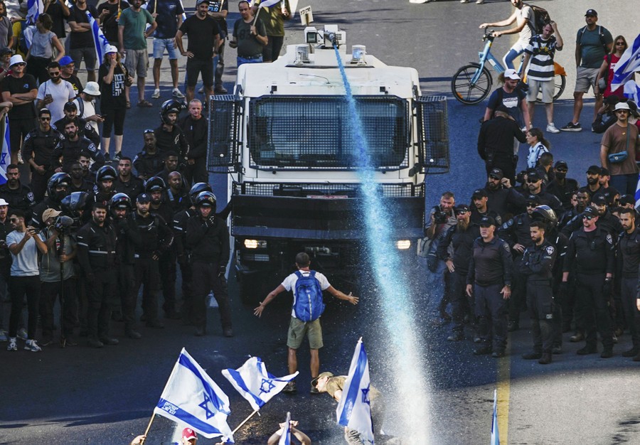 Riot police officers stand beside a vehicle-mounted water cannon that is spraying protesters.