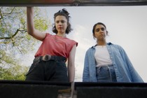 A still image of Margaret Qualley and Geraldine Viswanathan in “Drive-Away Dolls,” wearing puzzled expressions as they look into the trunk of a car. A gray-blue sky and green treetop are behind them.