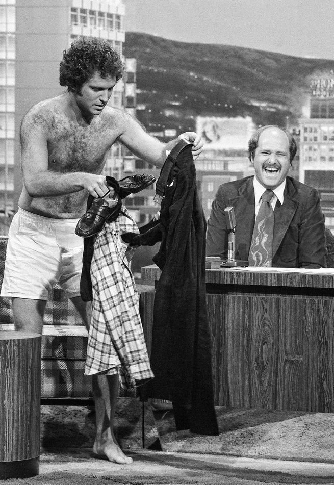 black-and-white photo of Brooks in boxers, holding pants, shirt, and belt in his hands, on a TV set with Reiner sitting behind host desk and laughing 