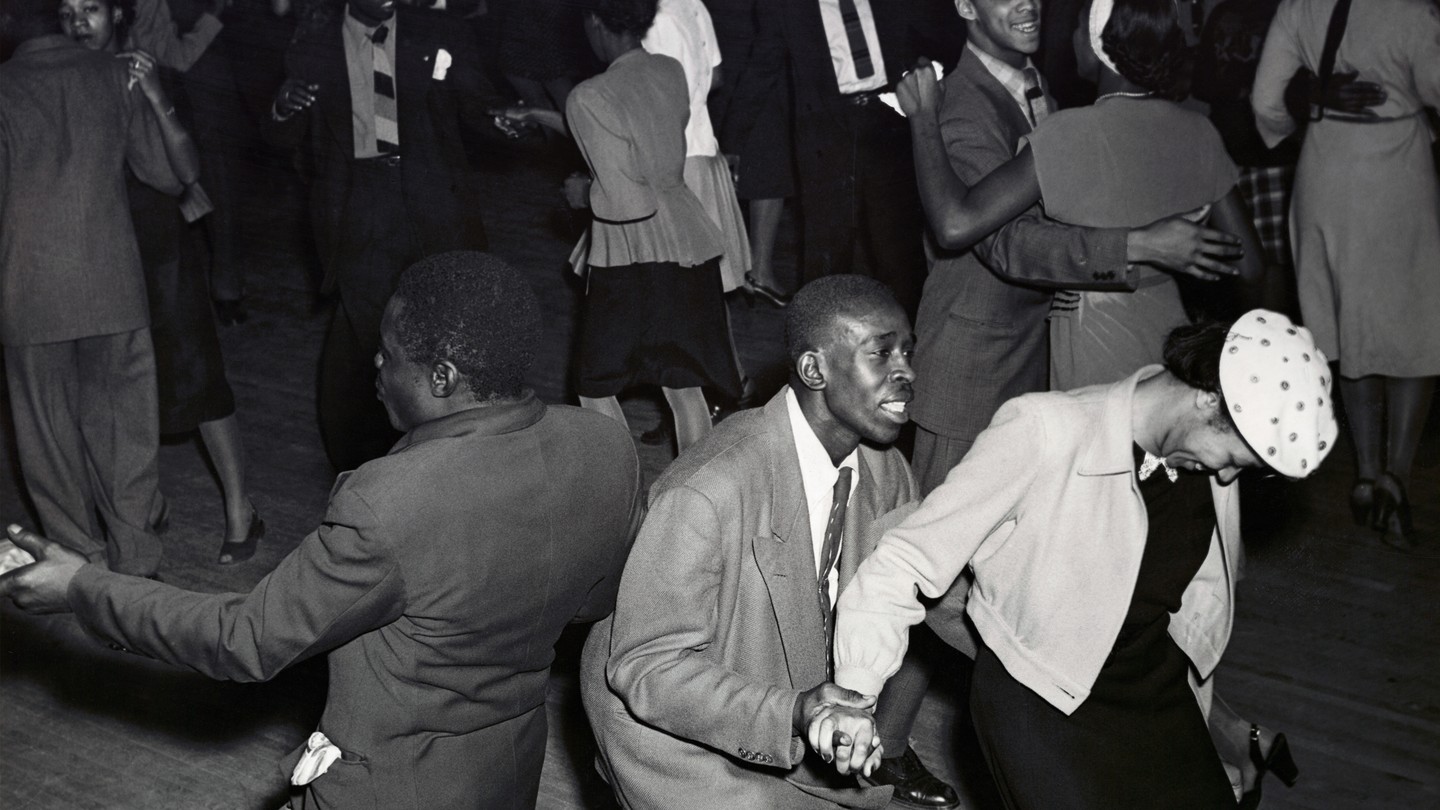 A black and white photo of two men and a woman dancing, on a crowded dance floor