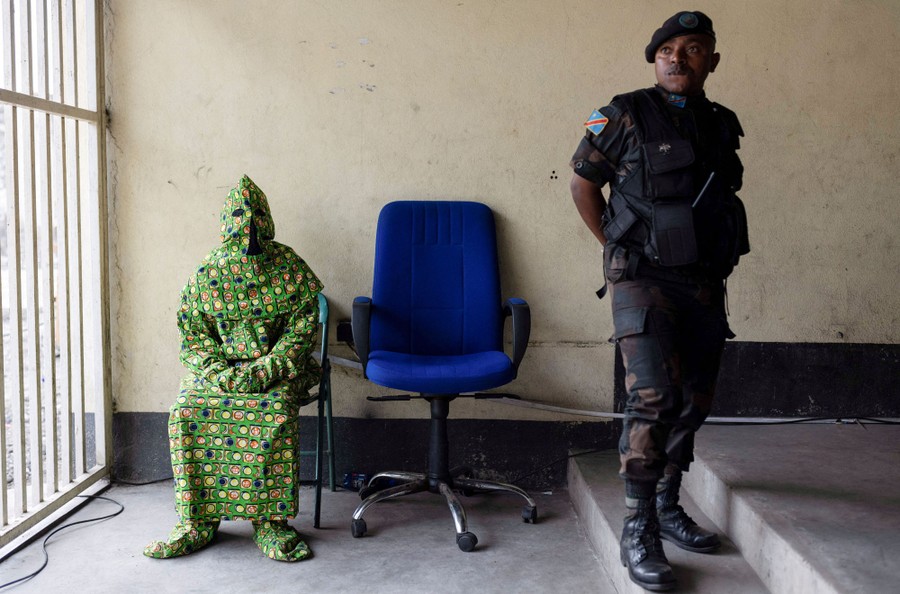 A person wears a patterned hood and full-body covering to disguise themselves as they sit in a chair near a police officer.