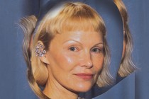 Pamela Anderson wearing an elaborate earring and matching blueish dress in front of a distorted outline of her image in front of a blue curtain