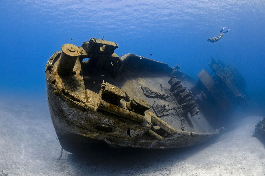 A shipwreck lies on the bottom of the sea, as a single person swims down toward it.