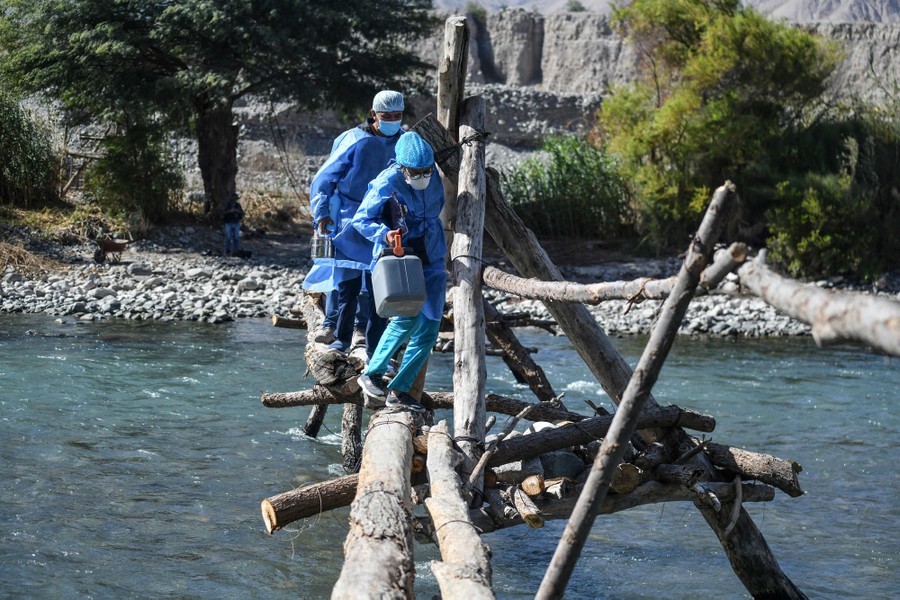 Health-care workers wearing protective gear cross a river on a rickety hand-built bridge.