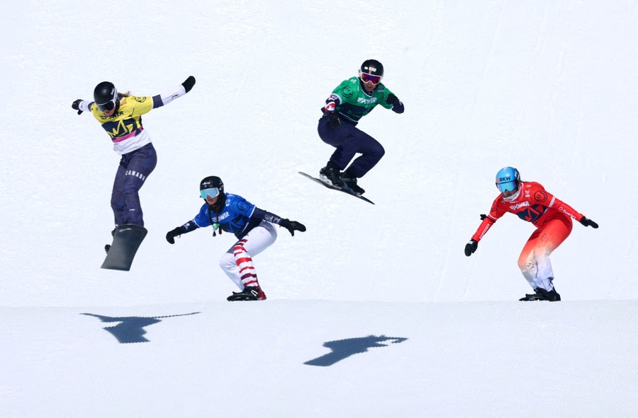 Four snowboarders race down a course, with two seen in mid-jump.