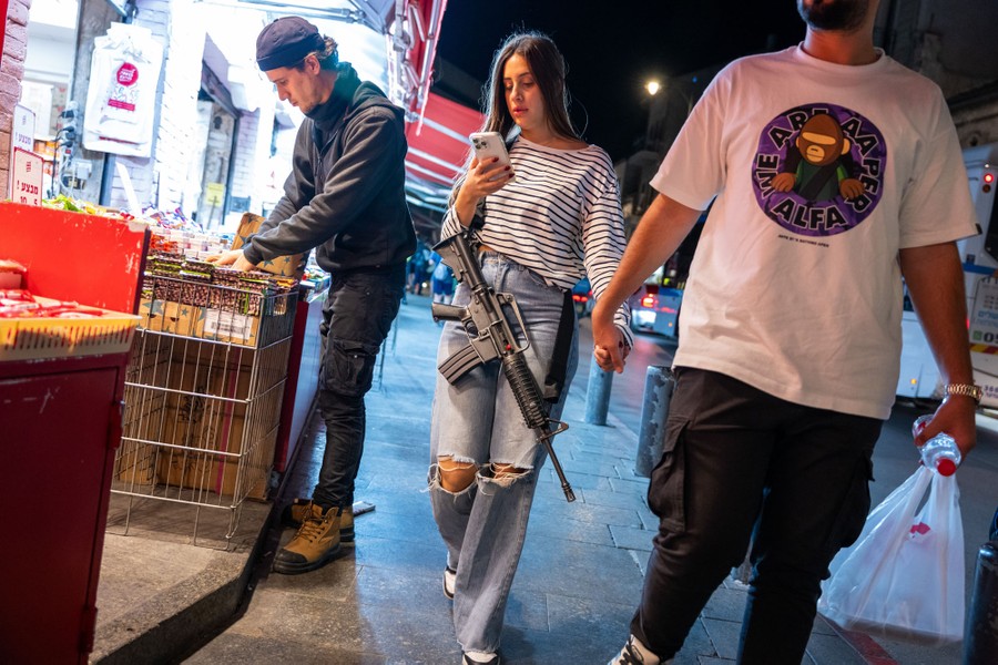 A couple walks on a sidewalk past a market, holding hands. The woman at center carries a rifle hanging from a strap over her shoulder, while using her smartphone.