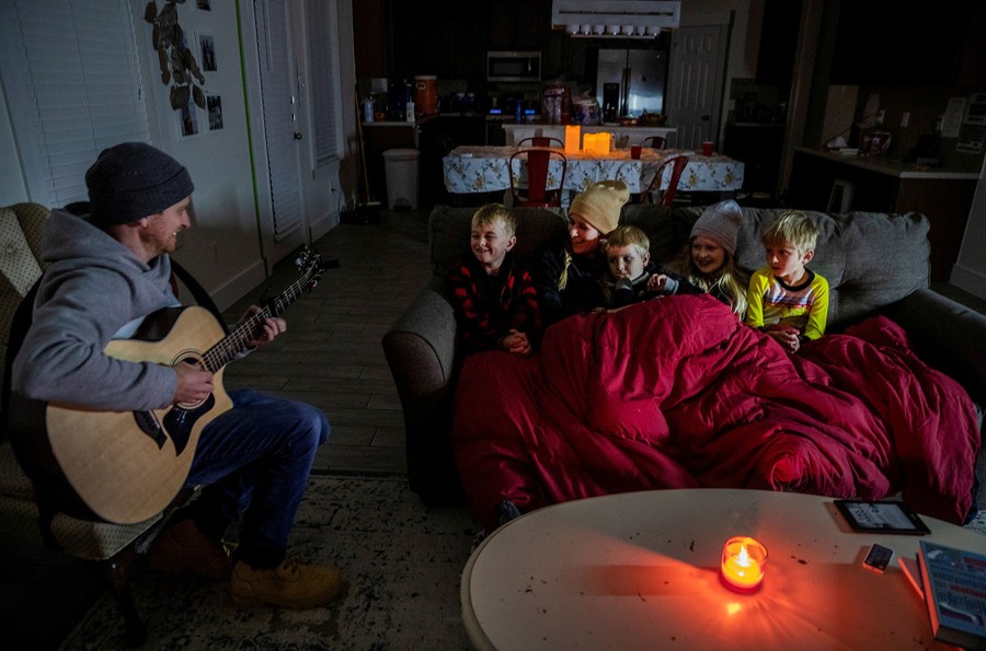 A man plays guitar in a living room for his family who are bundled up under a blanket on a couch.