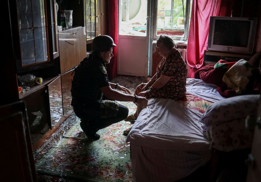 A police officer kneels down to help a seated woman in her apartment, which is strewn with broken glass.
