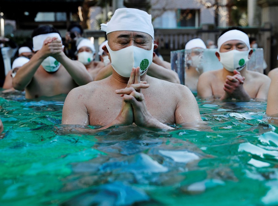 Several shirtless men wearing traditional head wraps and modern protective face masks sit together in prayer in cold green water.