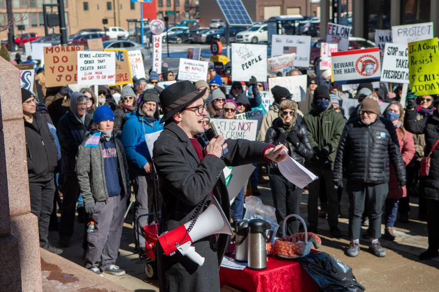A group of protesters holding signs gathers, listening to a person speaking into a megaphone.