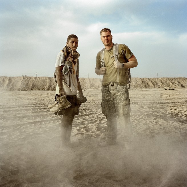 Two soldiers standing together, one holding boots in hands and one wearing backpack, with dust rising from ground