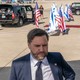 Photograph of J. D. Vance on an airport tarmac with U.S. flags, Israeli flags, black SUVs, and a red carpet behind him