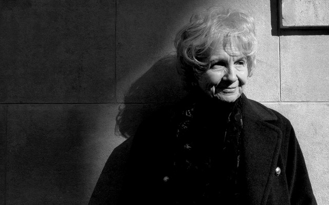 A black-and-white portrait of Alice Munro, standing against a wall and gazing to the right