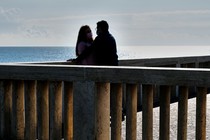 Two people wearing masks stand face-to-face on a dock, with a body of water in the background.