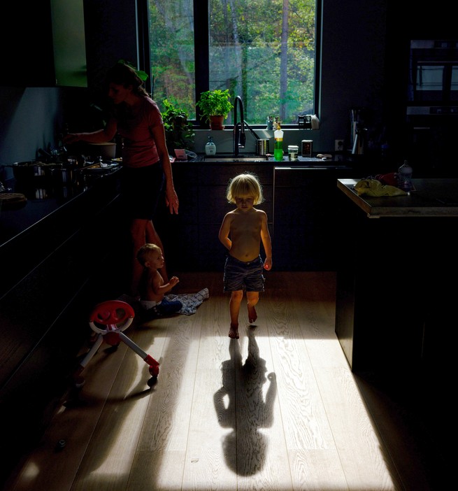 A photograph of a mother cooking in shadow. A child stands, well-lit, in the center of the kitchen.
