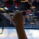 A pilot's hand extends up from the cockpit seat towards a ceiling full of switches. A pink manicured thumb nail is revealed and reaches towards a switch.