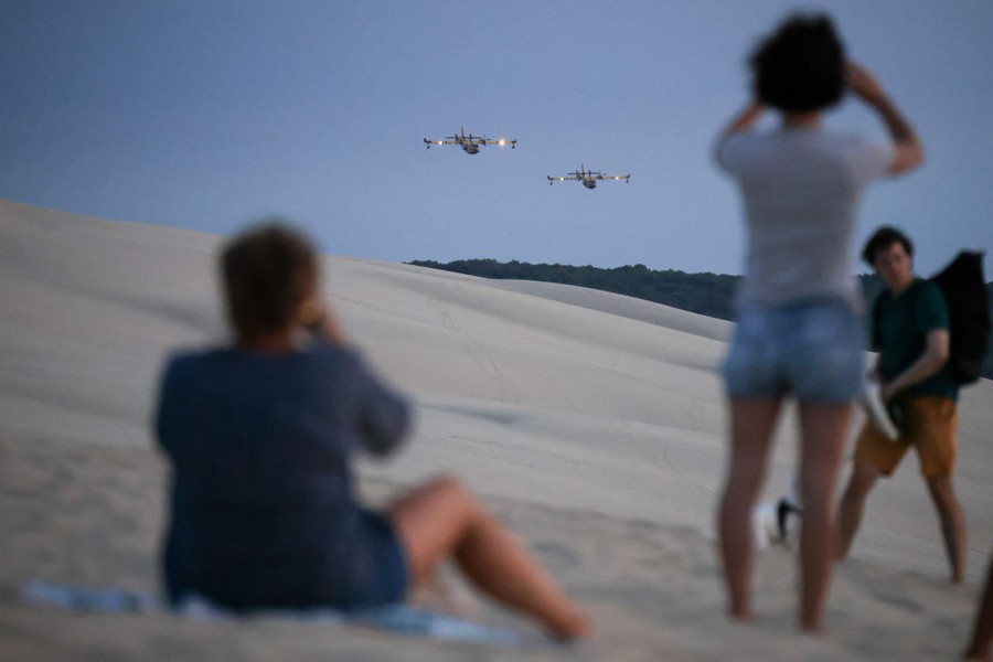 Two firefighting aircraft fly low over a broad sand dune and forest as several people watch in the foreground.