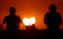 Two people look at a distant sunset, with part of the sun occluded by a partial eclipse.