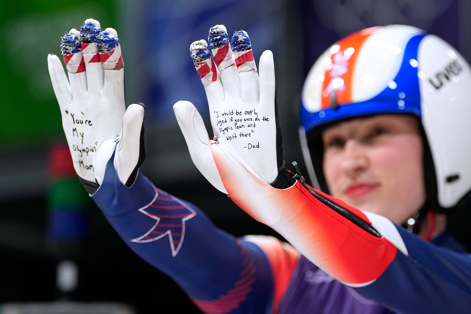 A luge athlete holds up their hands, showing encouraging messages from their parents written on the palms of their gloves.