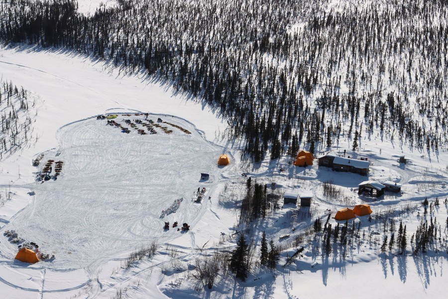 An aerial view of a cleared area of snow on a frozen body of water, with tents and support vehicles, as well as several resting dog teams.