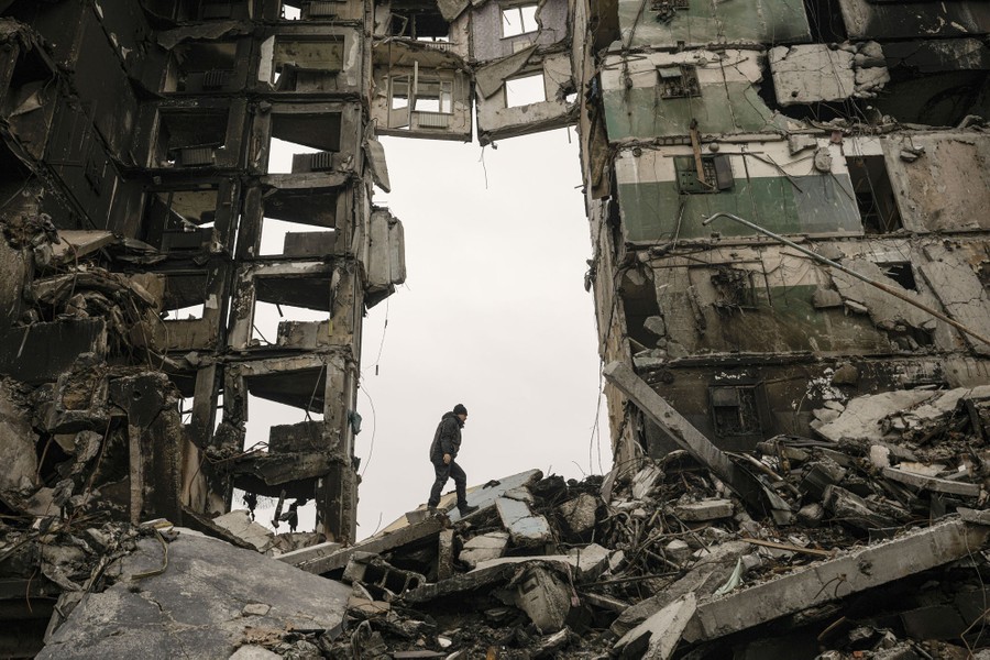 A person walks on rubble beside a heavily damaged apartment building that still partially stands.