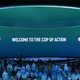 In a blue-lit room a blue and green ombre screen reads "welcome to the COP of action." A large circular blue light defines the ceiling.