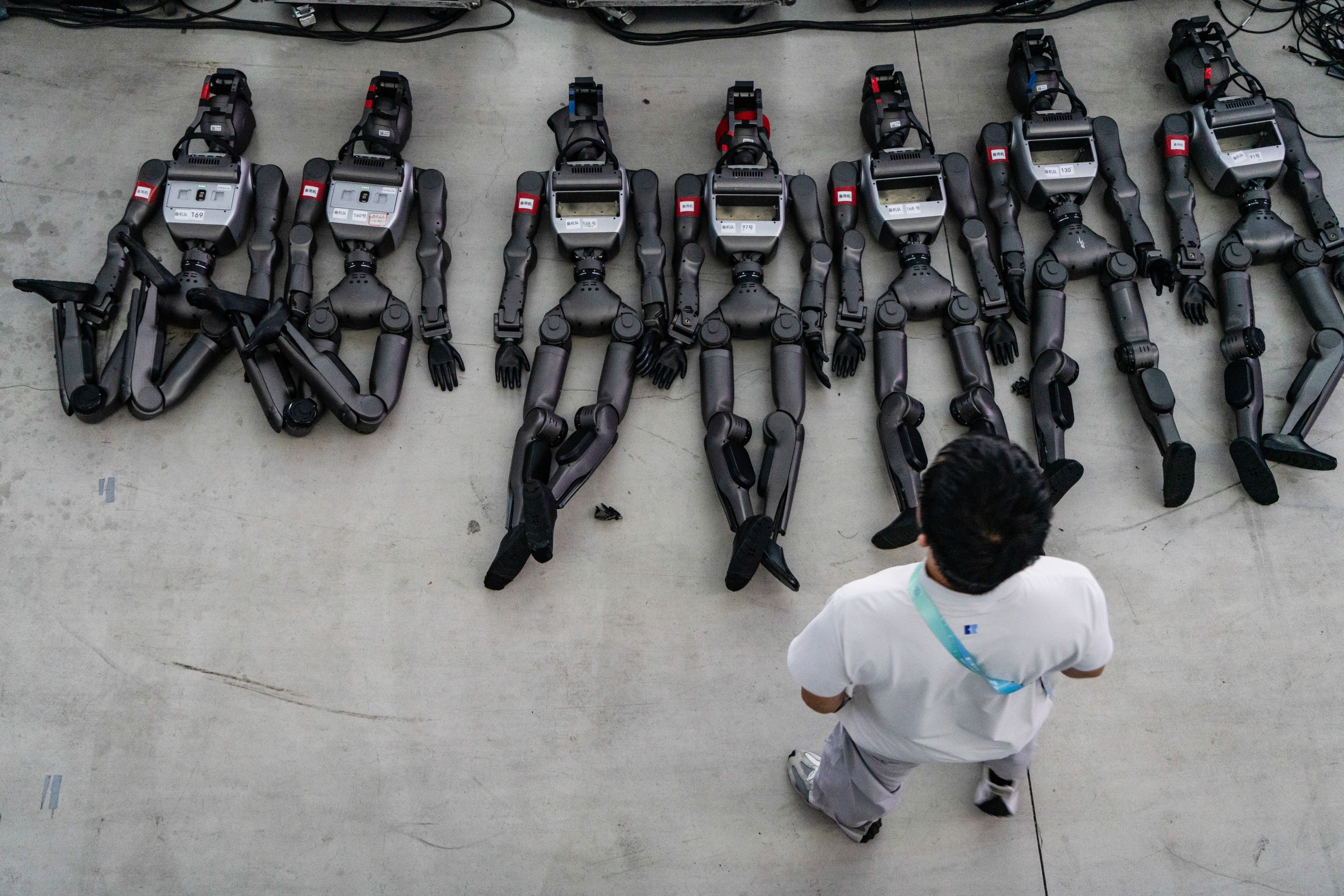 A person stands over seven humanoid robots that lie on concrete.