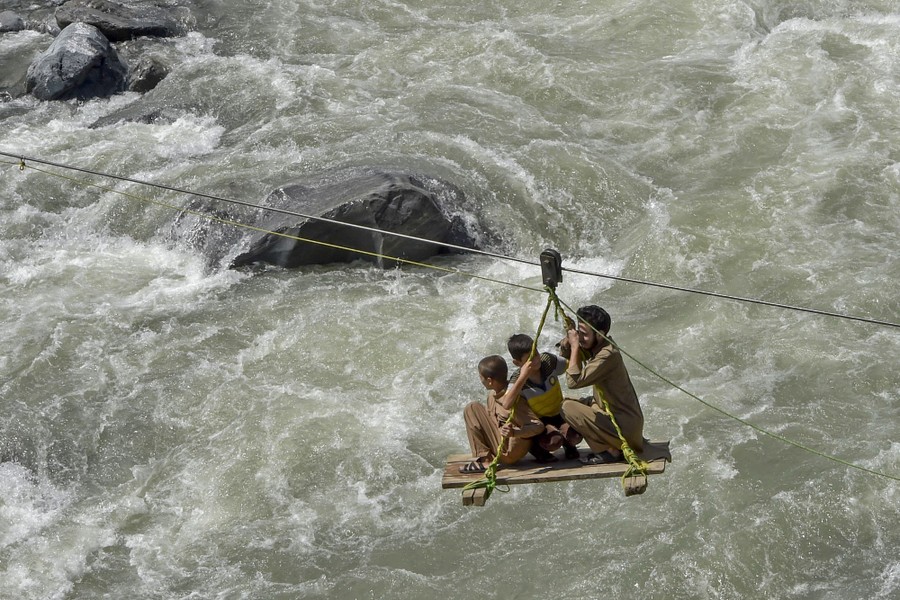 Three people ride on a plank suspended from a cable, crossing a fast-flowing river.