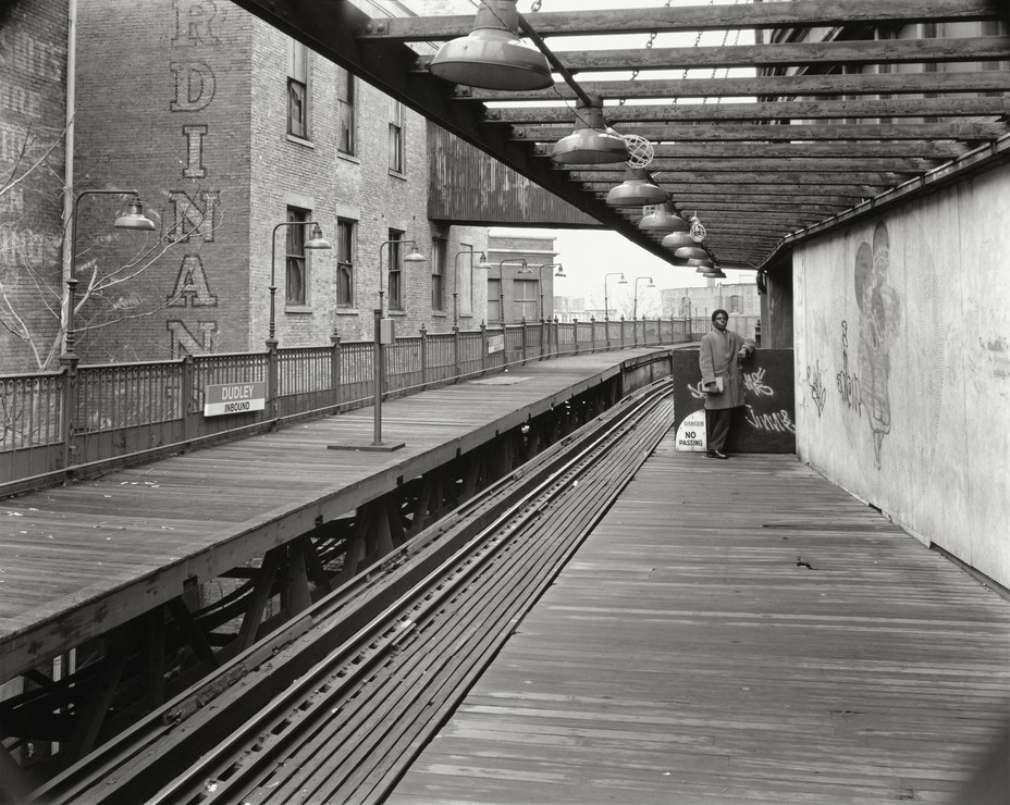 black-and-white photo of almost empty train-station platform with person standing at far end in coat