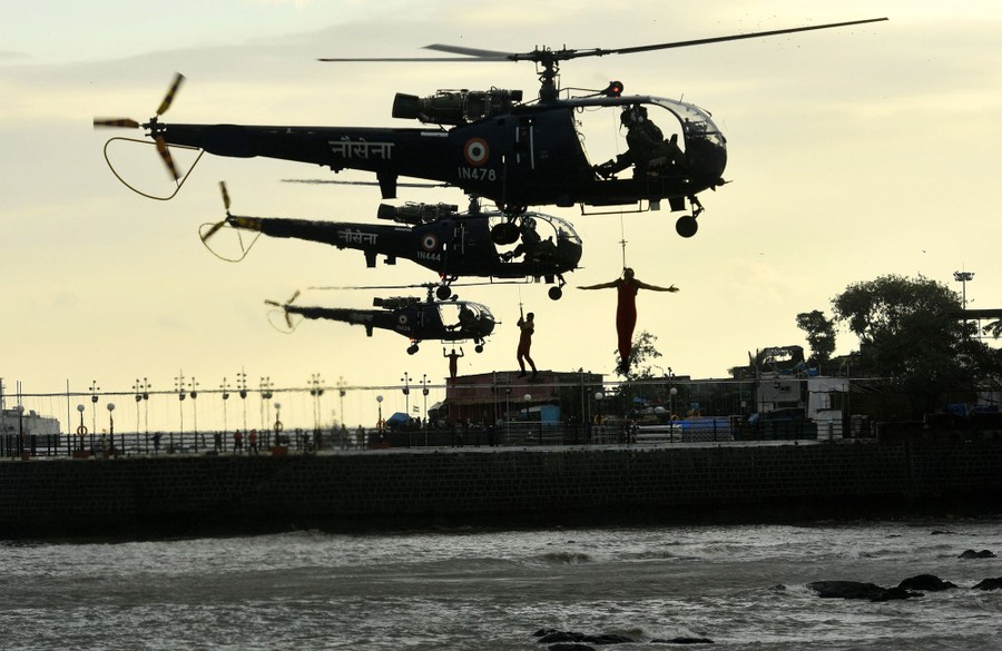 Three helicopters hover near the water's surface, dangling one man each from a rope below.