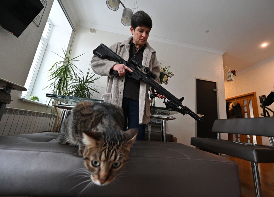 A woman stands in an apartment, holding a long rifle, beside a cat.