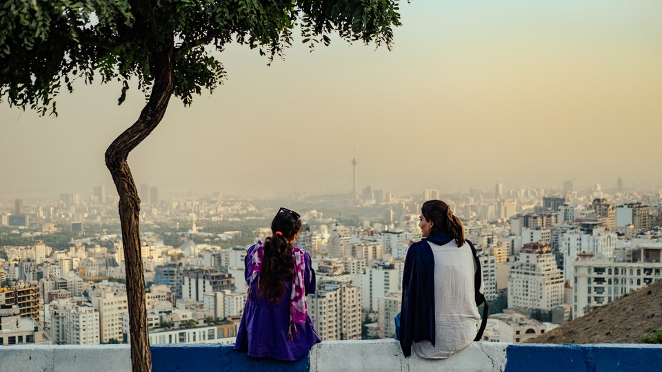Two women with uncovered hair sit on a wall overlooking the city of Tehran.