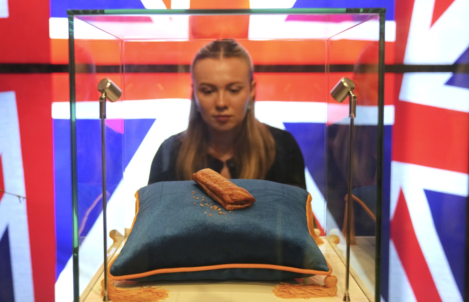 A person looks at a sausage roll that has been placed on a pillow under a glass display case, backdropped by the flag of the United Kingdom.