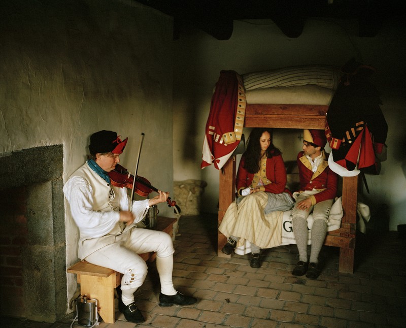 photo of room inside the English quarters of Fort Ticonderoga, where reenactors play a soldier playing the fiddle and two younger English residents sitting and talking on a wooden bed