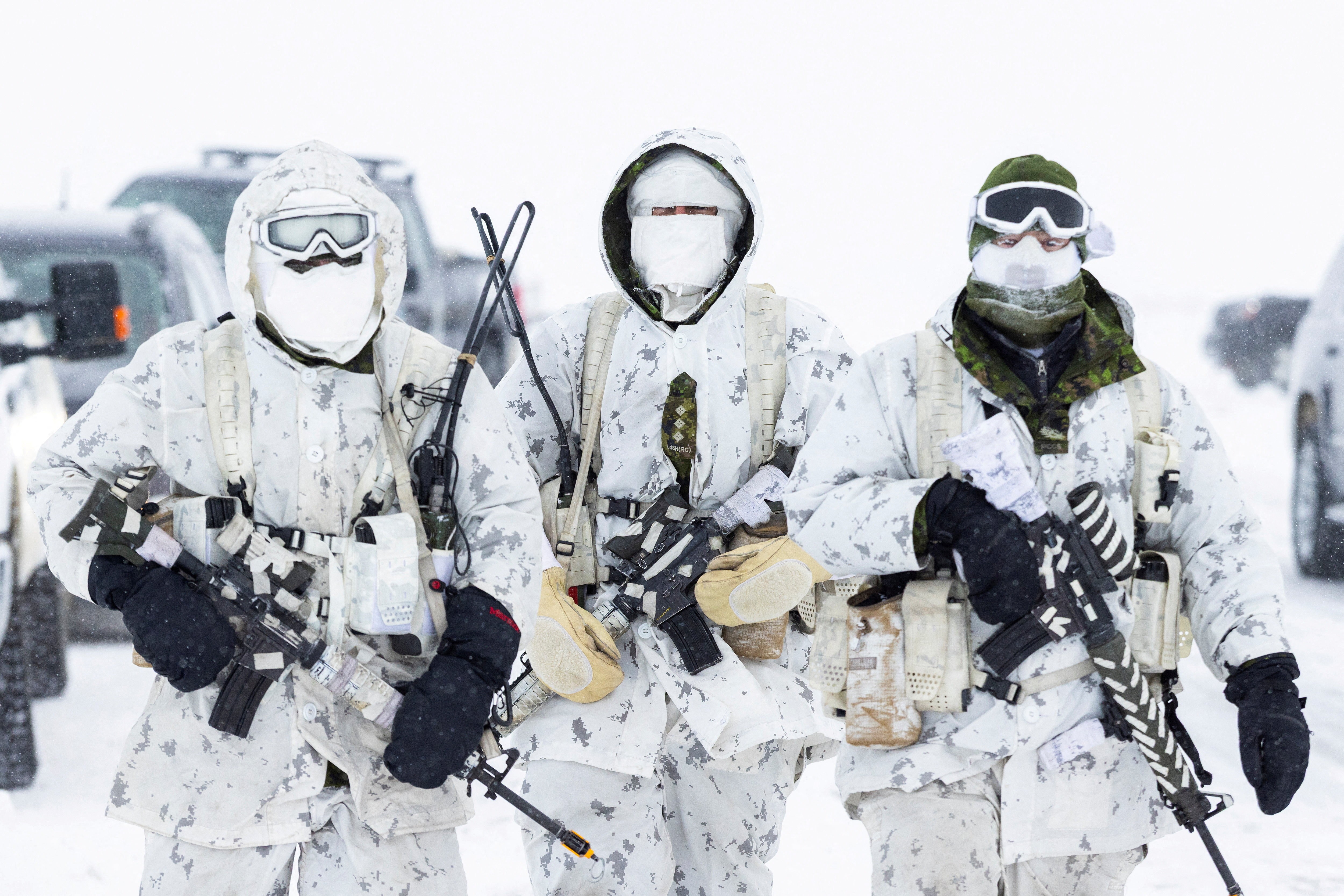 Three soldiers walk down a road in a snow-covered area. All wear white camouflage uniforms and carry weapons.