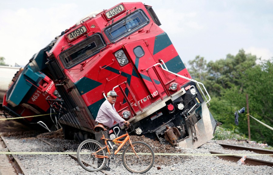 A man walks a bicycle in front of a derailed locomotive.