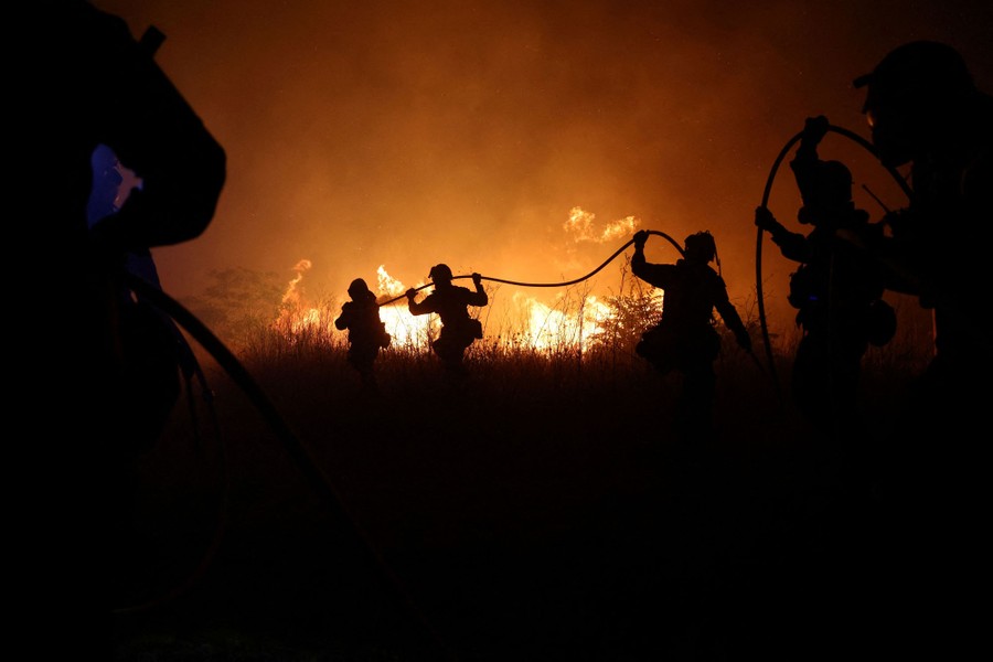 A half-dozen firefighters wrangle a hose, moving toward wildfire flames at night.