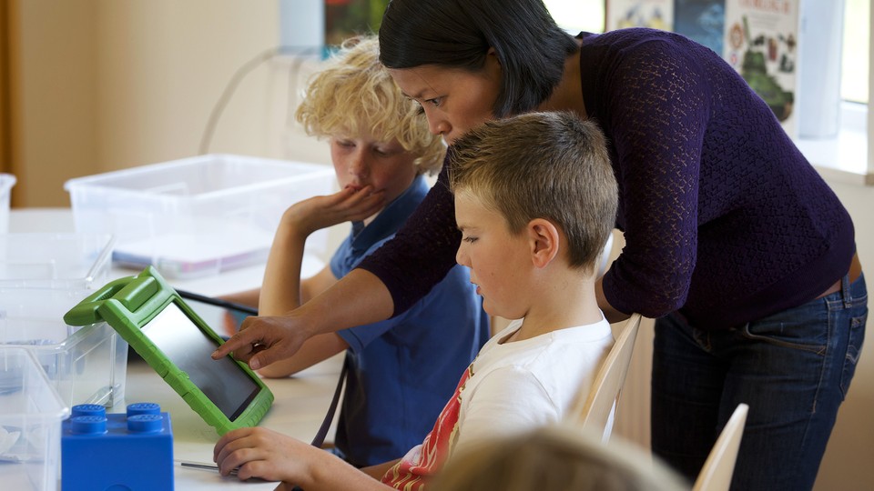 A teacher stands next to two students and looks at something on an iPad with them.