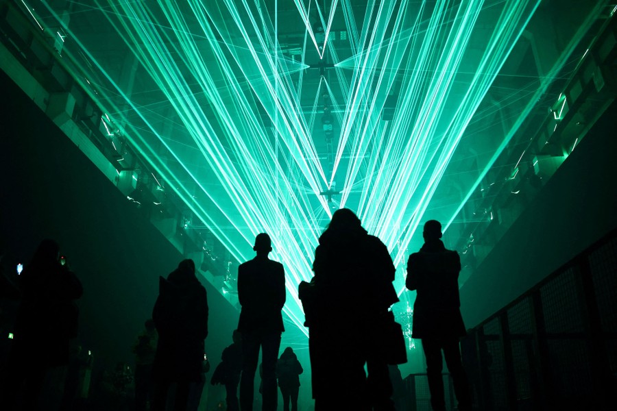 People stand and look up at many green rays of light shining along the ceiling of a tall room.